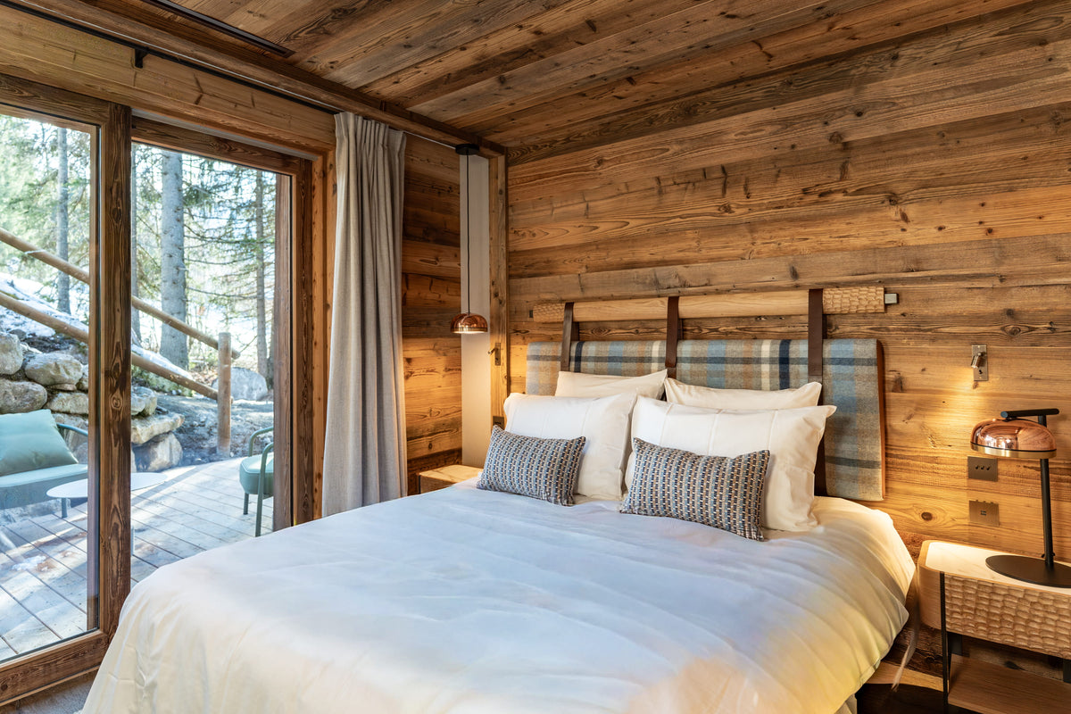A cozy bedroom with a neatly made bed dressed in white bedding and patterned pillows, set against rustic wood-paneled walls and a large window looking out onto a forest deck.