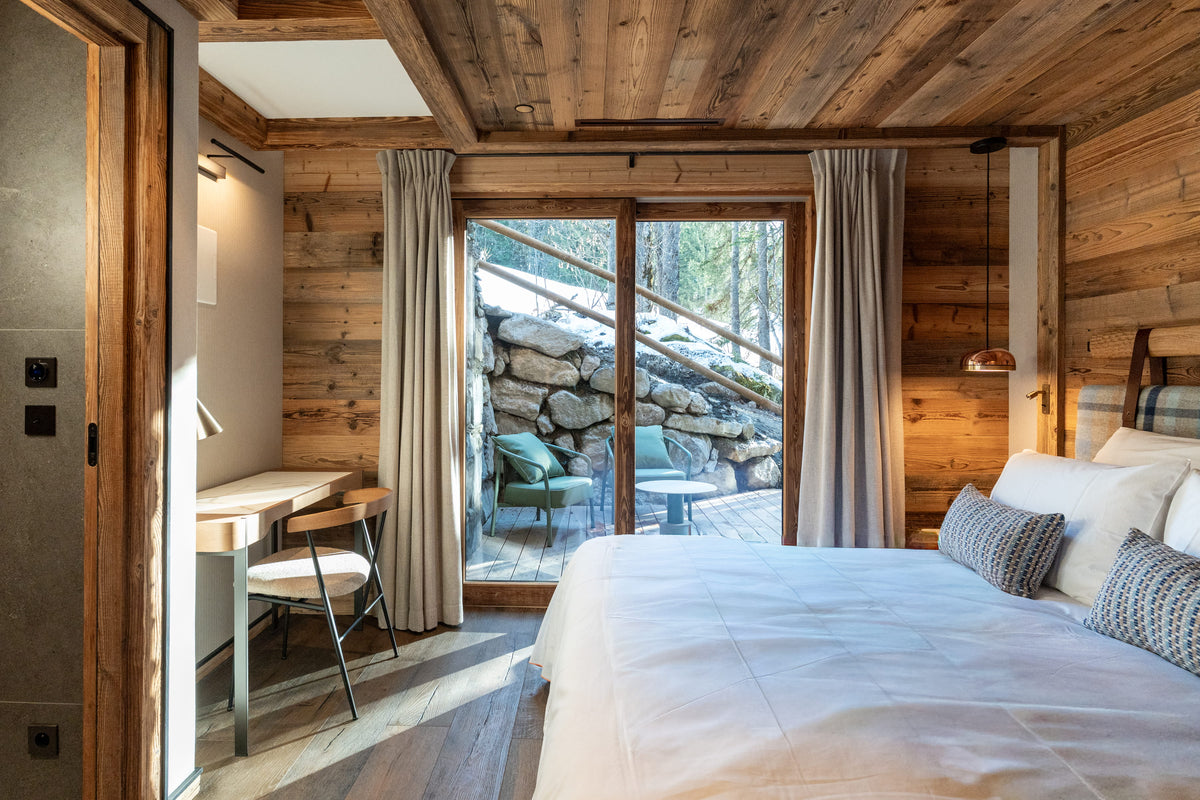 A rustic mountain chalet bedroom featuring a large bed with white linens, a wooden desk and chair, and a large glass sliding door looking out to a stone patio and forest.