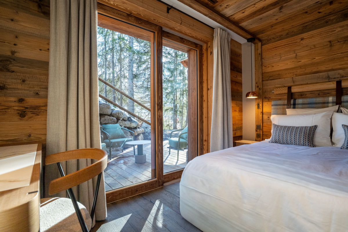 A cozy bedroom interior with a large bed dressed in white linens, wood-paneled walls and ceiling, beige curtains, and floor-to-ceiling glass doors opening to a forest view and outdoor patio.