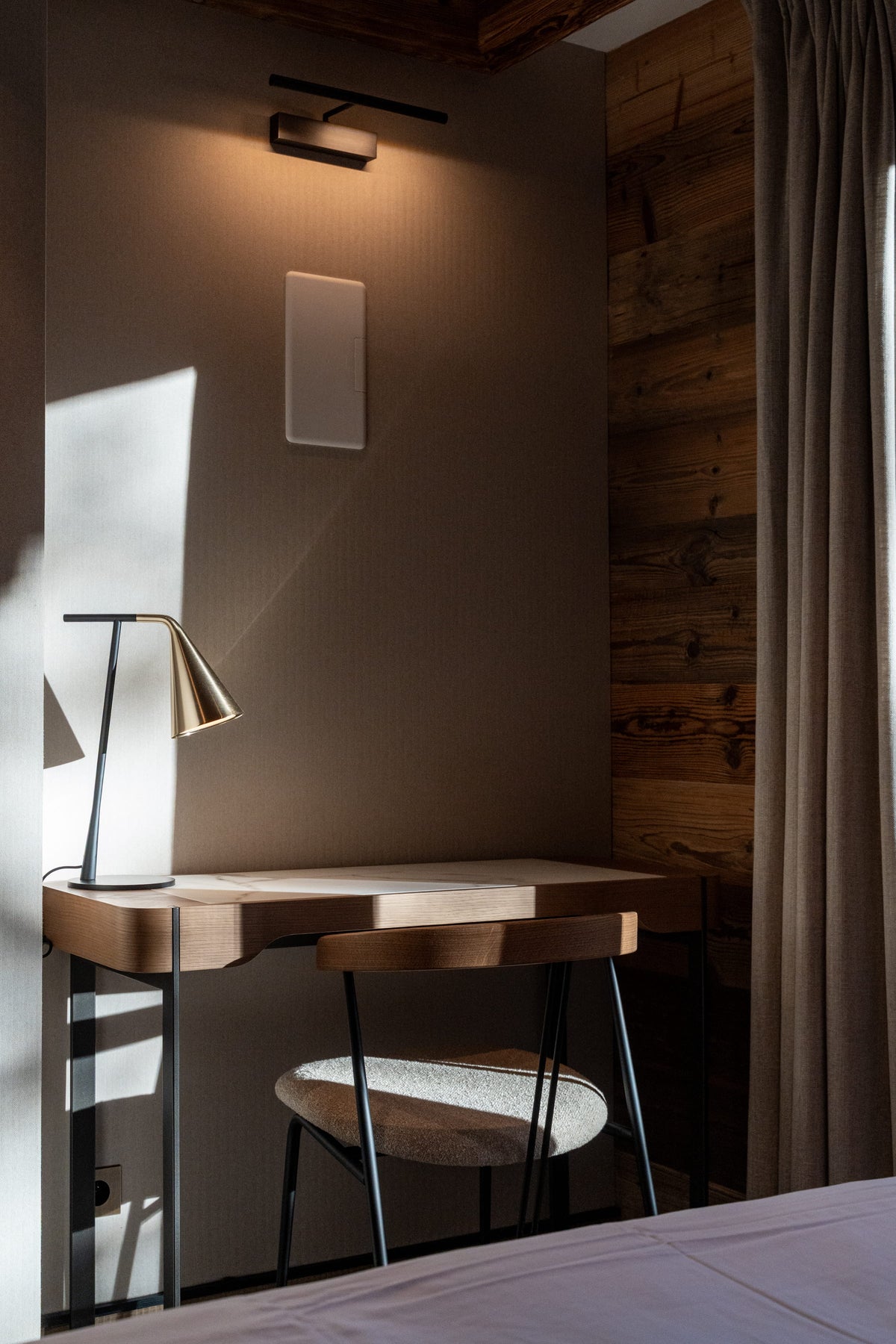 A modern minimalist bedroom corner featuring a wooden desk and matching chair with a sleek black metal lamp, set against a warm brown wall and wood-paneled side wall with a curtain.