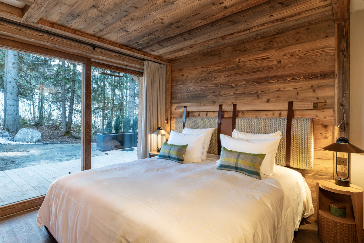 A cozy bedroom with a large bed dressed in white bedding and two green plaid accent pillows, set against rustic wood-paneled walls and a wooden ceiling. Large windows on the left offer a view of a snowy forest outside.