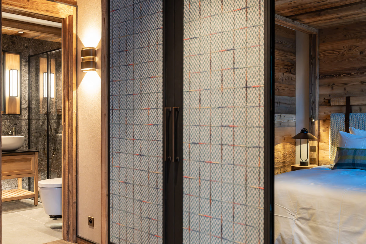 A modern bedroom interior with a sliding glass door featuring a geometric grid pattern, separating the bedroom from an adjacent bathroom and log-cabin style bedroom area.