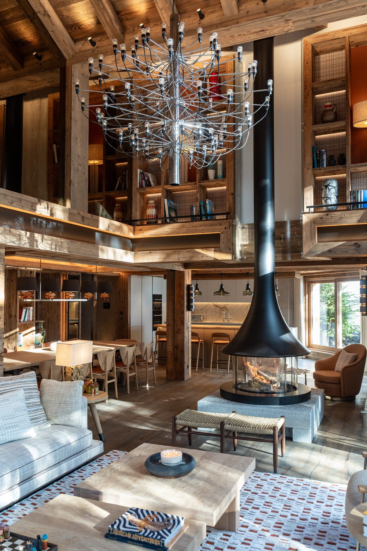 A large, rustic chalet-style living room with high wooden ceilings, a massive multi-arm chandelier, a central black flue chimney, and a modern fireplace. The room features a light blue sofa, a low wooden coffee table, patterned tile flooring, and built-in wooden bookshelves.