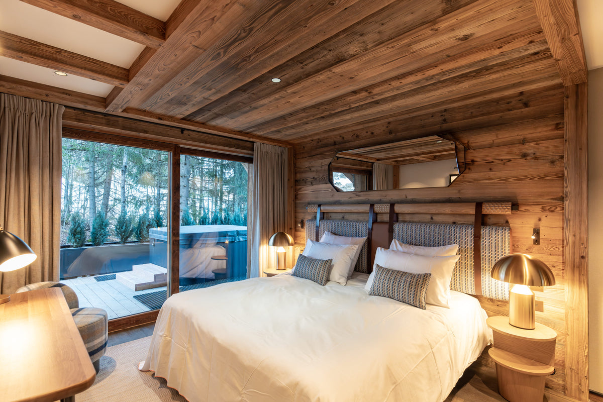 A cozy bedroom with a large bed dressed in white linens and striped pillows, set in a rustic wooden cabin with wood-paneled walls and ceiling, large windows showing an outdoor deck and forest view, and warm bedside lamps.