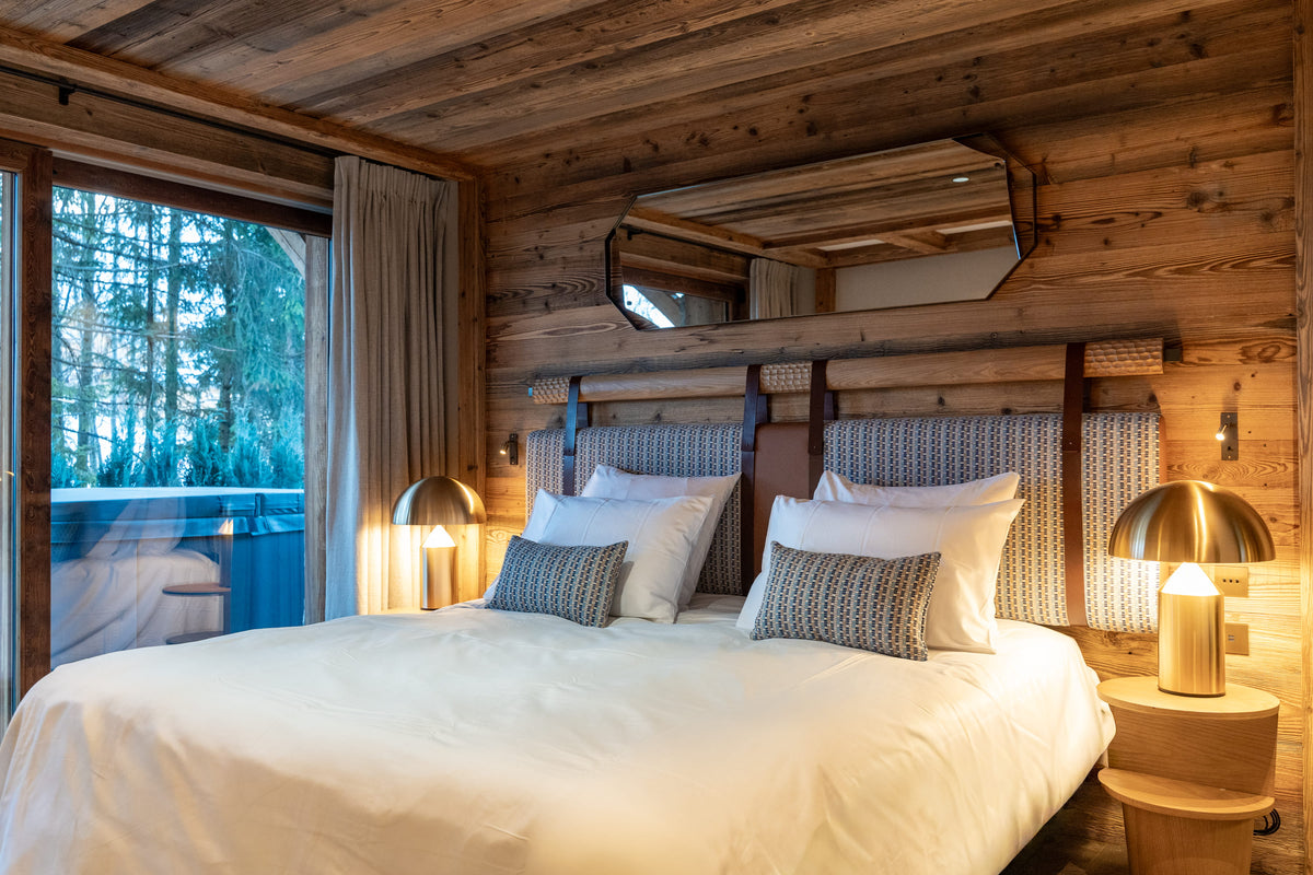 A cozy bedroom with a neatly made bed, white linens, patterned pillows, wooden walls, bedside tables, and lamps. A large window on the left shows a forest view outside.