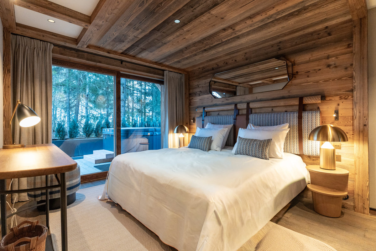A luxury bedroom with a large bed dressed in white linens, set against rustic wood-paneled walls and ceiling. Two modern bedside tables with warm brass lamps sit on either side of the bed, and a large window on the left looks out onto a snowy forest.