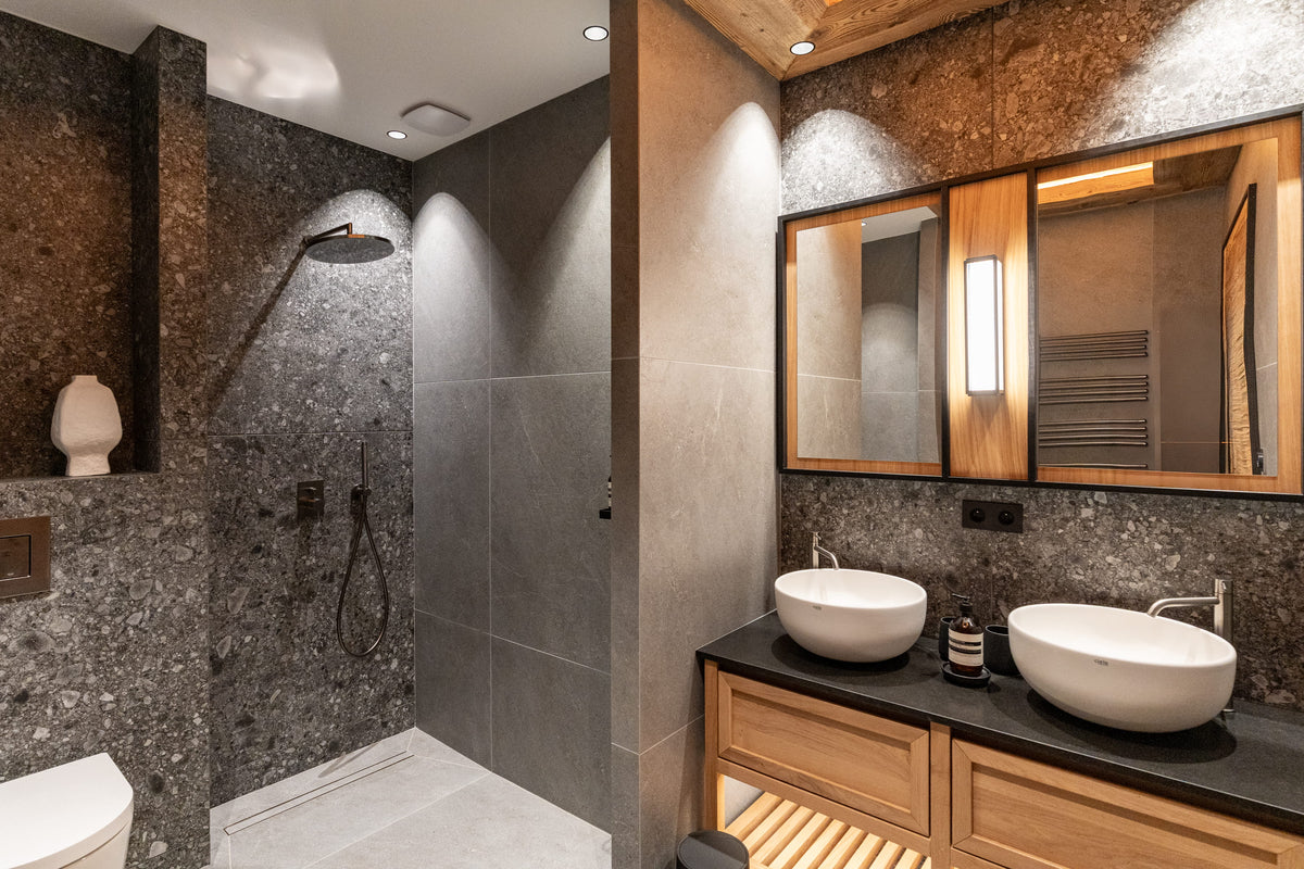 Modern luxury bathroom interior featuring a walk-in shower with dark stone walls, a double vessel sink vanity with wood cabinets and mirrors, and warm ambient lighting.