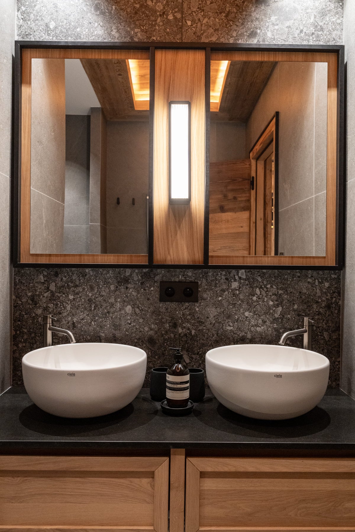 A modern double bathroom vanity with two white vessel sinks on a dark countertop, a wood cabinet base, and a large mirrored wall cabinet with warm lighting above.