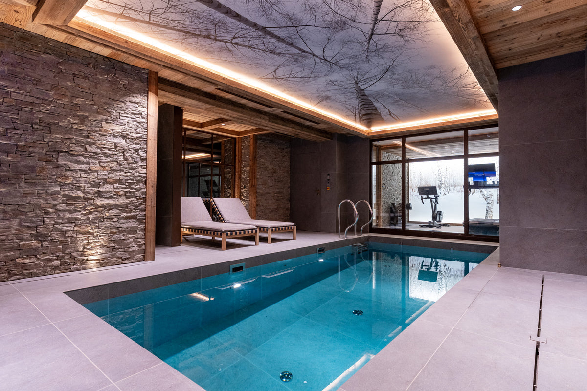 An indoor heated pool with bright blue water, surrounded by light stone tile decks. One wall features a rustic stacked stone facade with warm under-cabinet lighting, while the ceiling has a large decorative tree branch print with integrated LED lighting.