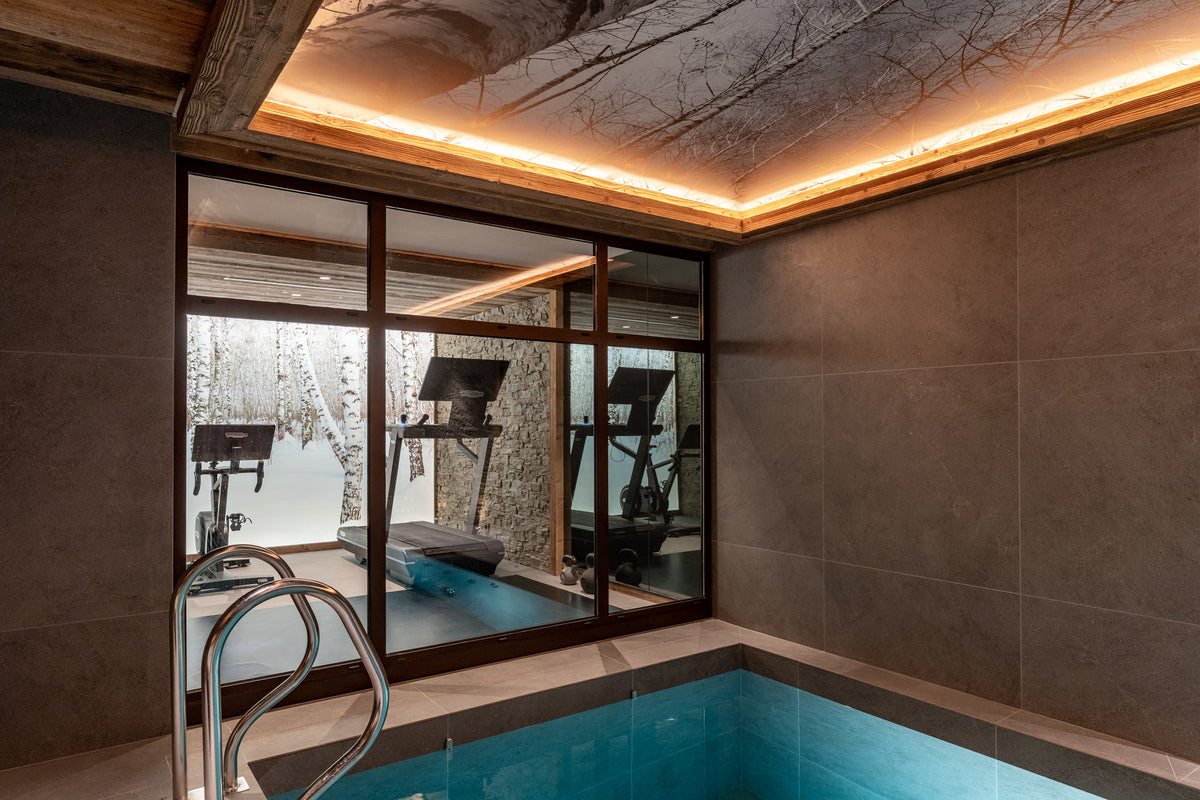An indoor pool area with a rectangular blue pool in the foreground, large gray stone walls, dark wood-framed glass doors, and exercise equipment visible through the glass. A metal pool ladder is in the lower left.