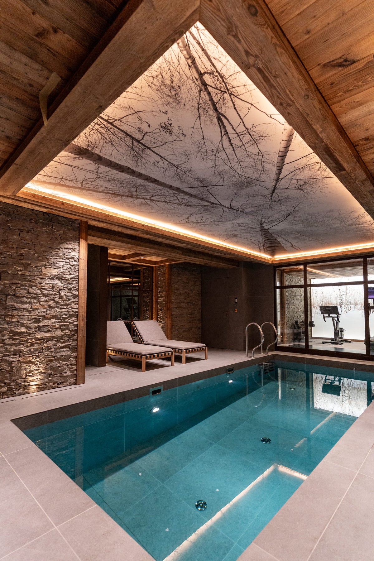 An indoor heated swimming pool in a luxury chalet with a stone wall, wood ceiling beams, and a tree-patterned ceiling light, featuring two lounge chairs by the pool.