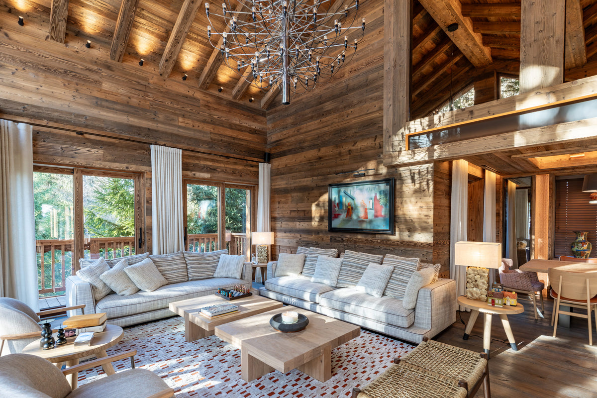 A luxury mountain chalet living room featuring a large white sectional sofa, wooden coffee tables, a patterned rug, and a rustic wood-paneled ceiling with a large chandelier.