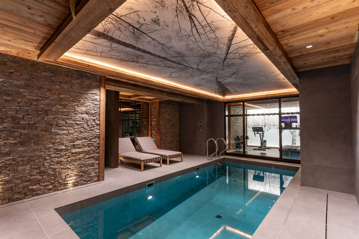 An indoor luxury pool area with a rectangular blue swimming pool, stone wall, wood ceiling beams, built-in LED lighting, and two lounge chairs near the poolside.