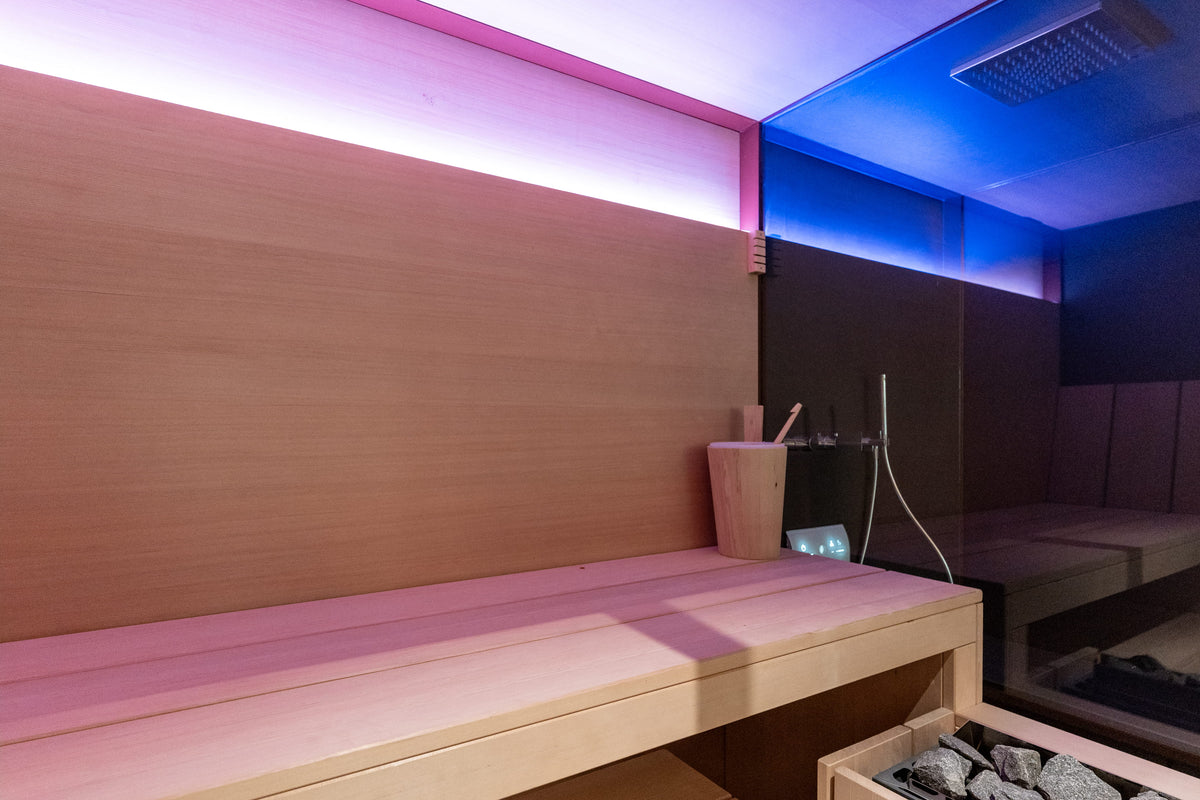 A modern wooden sauna room interior with a light wood bench, ambient pink and blue LED lighting, and a glass shower panel.
