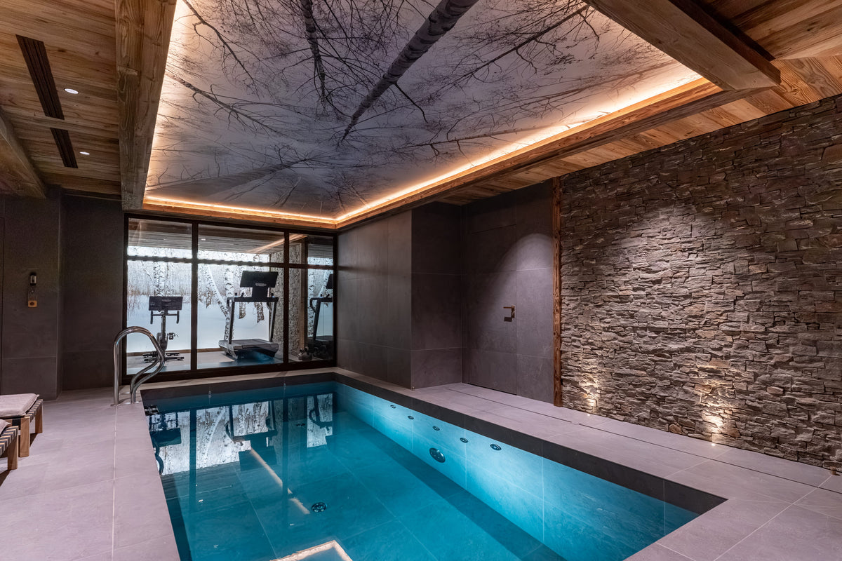 An indoor swimming pool in a luxury spa or resort setting, featuring a stone wall, wood ceiling with integrated lighting, and a glass window looking out onto a snowy outdoor scene. Exercise equipment is visible in the gym area beyond the window.