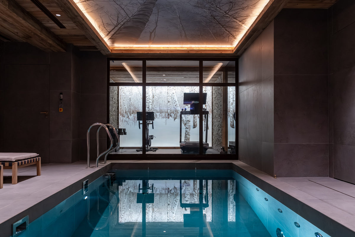 An indoor heated swimming pool with clear blue water, surrounded by dark stone walls and a modern glass-walled fitness area in the background.