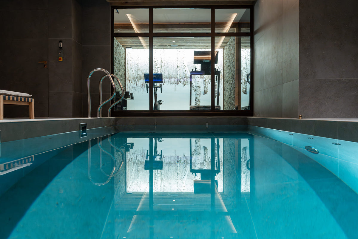 An indoor swimming pool with clear turquoise water, dark tiled walls, a metal ladder, and large glass doors reflecting the room behind it.