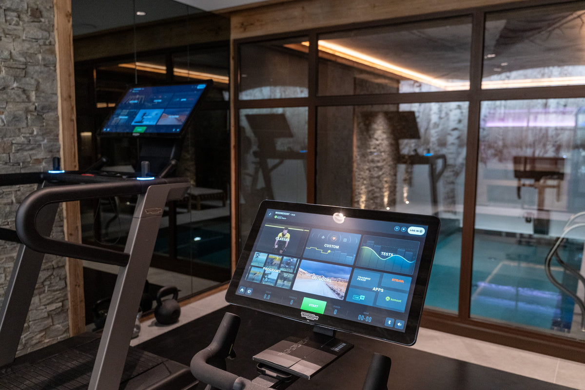 A modern treadmill in a fitness center with a large touch screen display on the console, set against large windows overlooking a swimming pool.