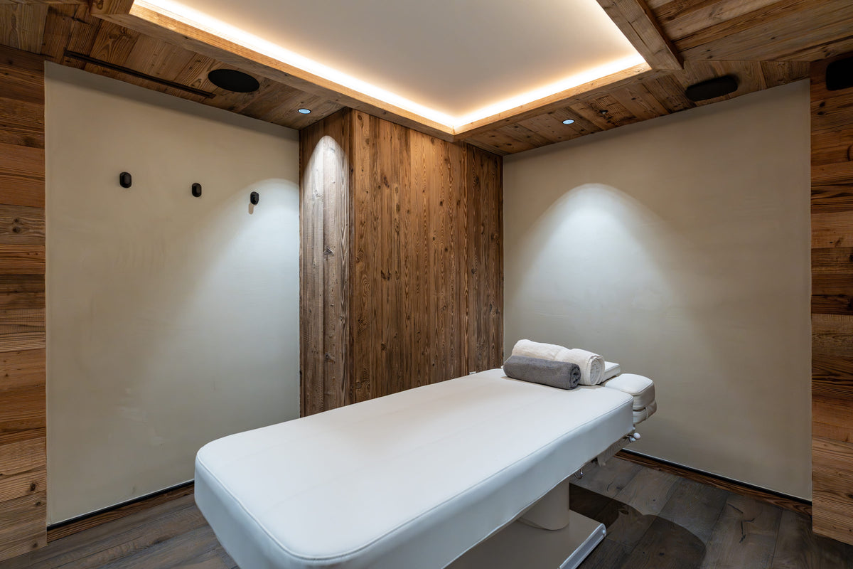 A white spa treatment table with a folded gray towel, set in a rustic room with wood-paneled walls and ceiling, recessed lighting, and a warm ambient glow.
