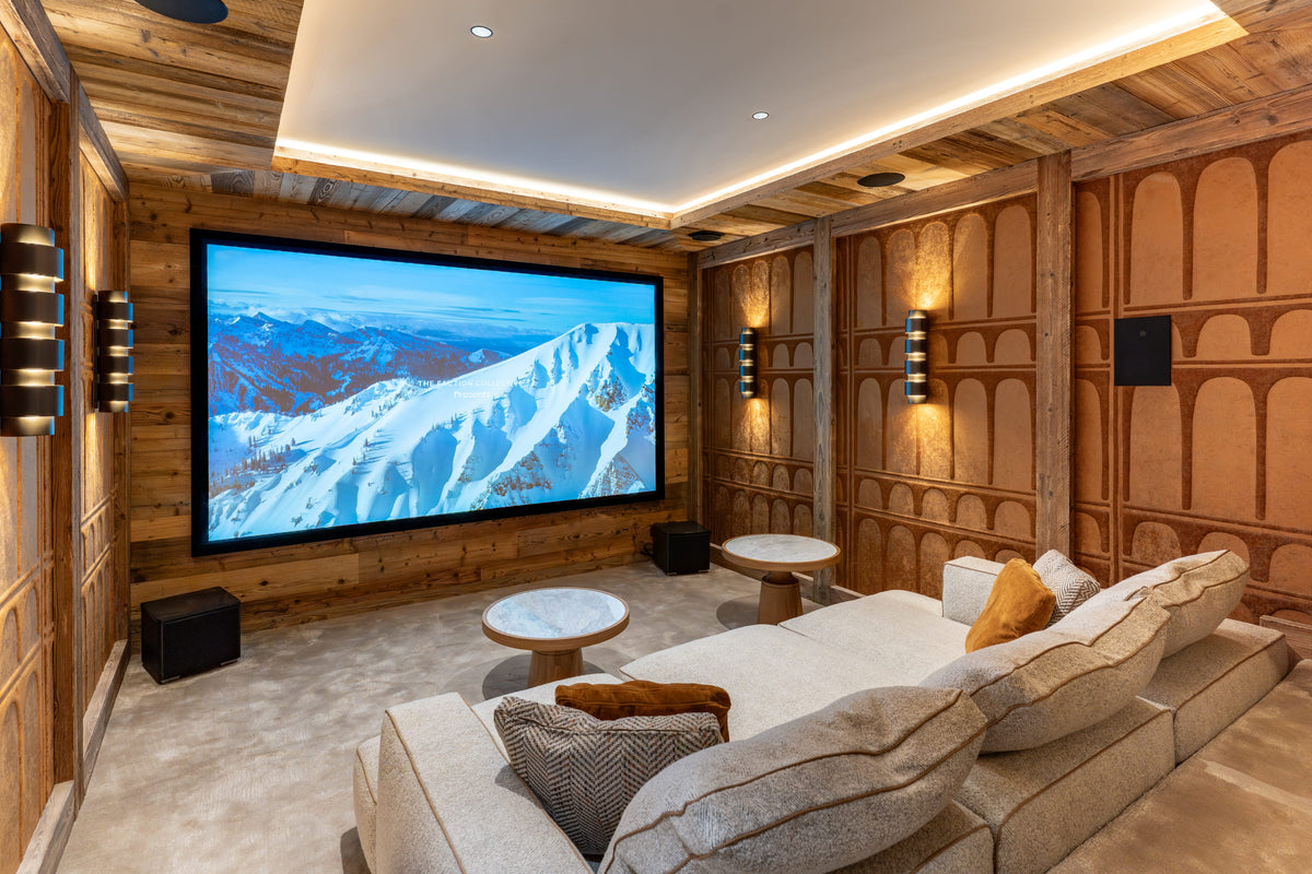 A luxury home theater with wood-paneled walls, a large screen showing a snowy mountain landscape, a plush beige sectional sofa, and ambient lighting.
