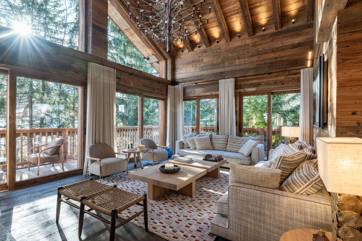 A bright, sunlit mountain chalet living room with high wood ceilings, large windows, and views of the forest outside. The room is furnished with a light beige sofa, matching armchair, patterned rug, wooden coffee table, and warm ambient lighting.