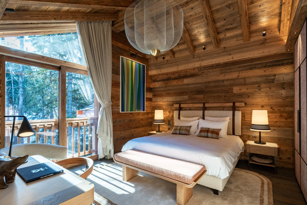 A cozy, rustic mountain-style bedroom with wood-paneled walls, a large bed with white linens, bedside tables and lamps, a desk in the foreground, and a balcony view visible through the windows.