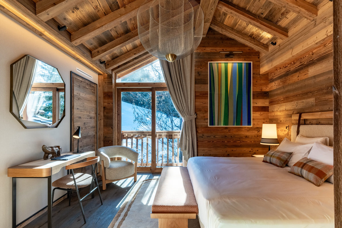 A luxury ski chalet bedroom featuring rustic wood-paneled walls and ceiling, a large bed with white linens, a wooden desk with a chair, and a colorful abstract painting on the wall.