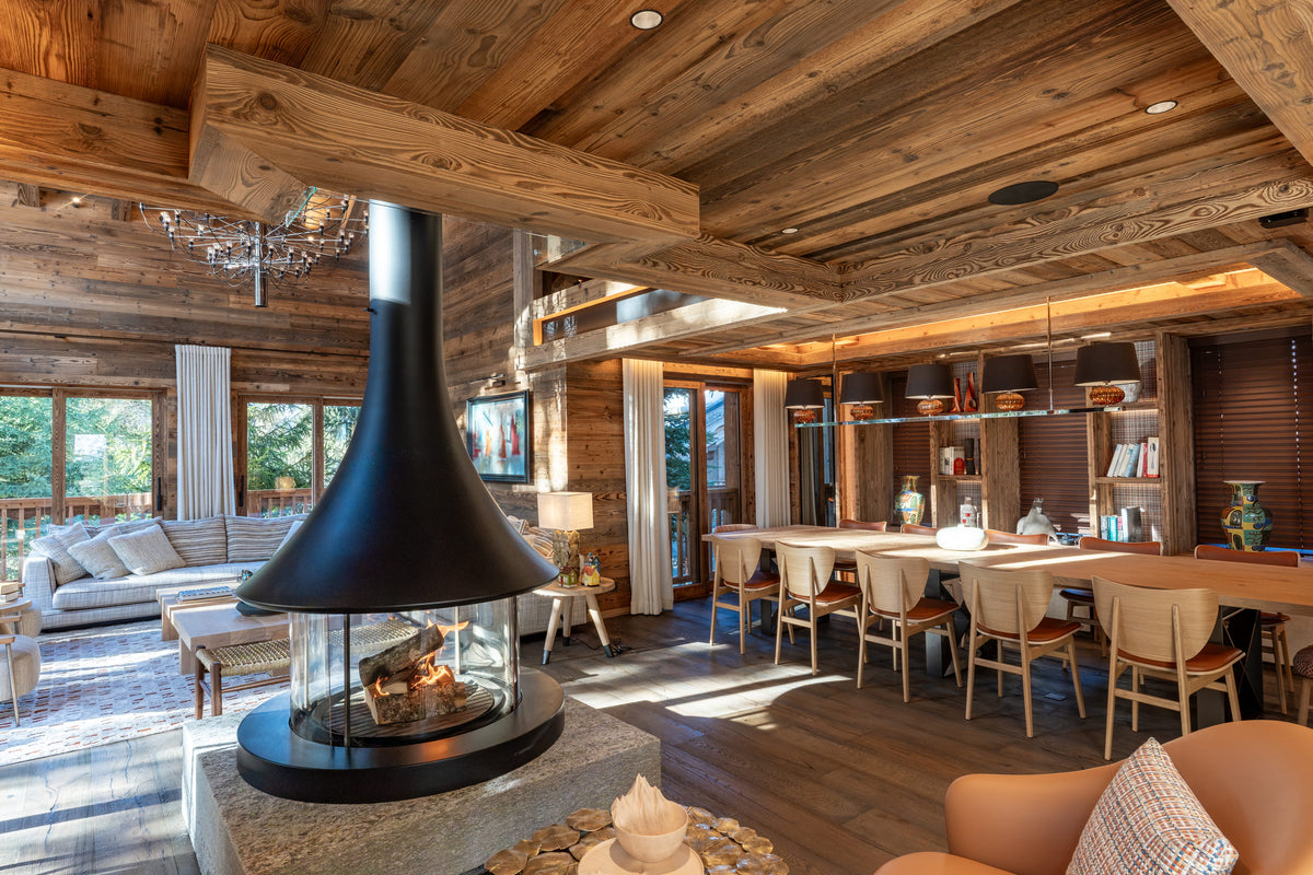 A modern black cone-shaped fireplace with a glass wind guard is featured in a rustic, wood-paneled chalet living room with a large dining area in the background.