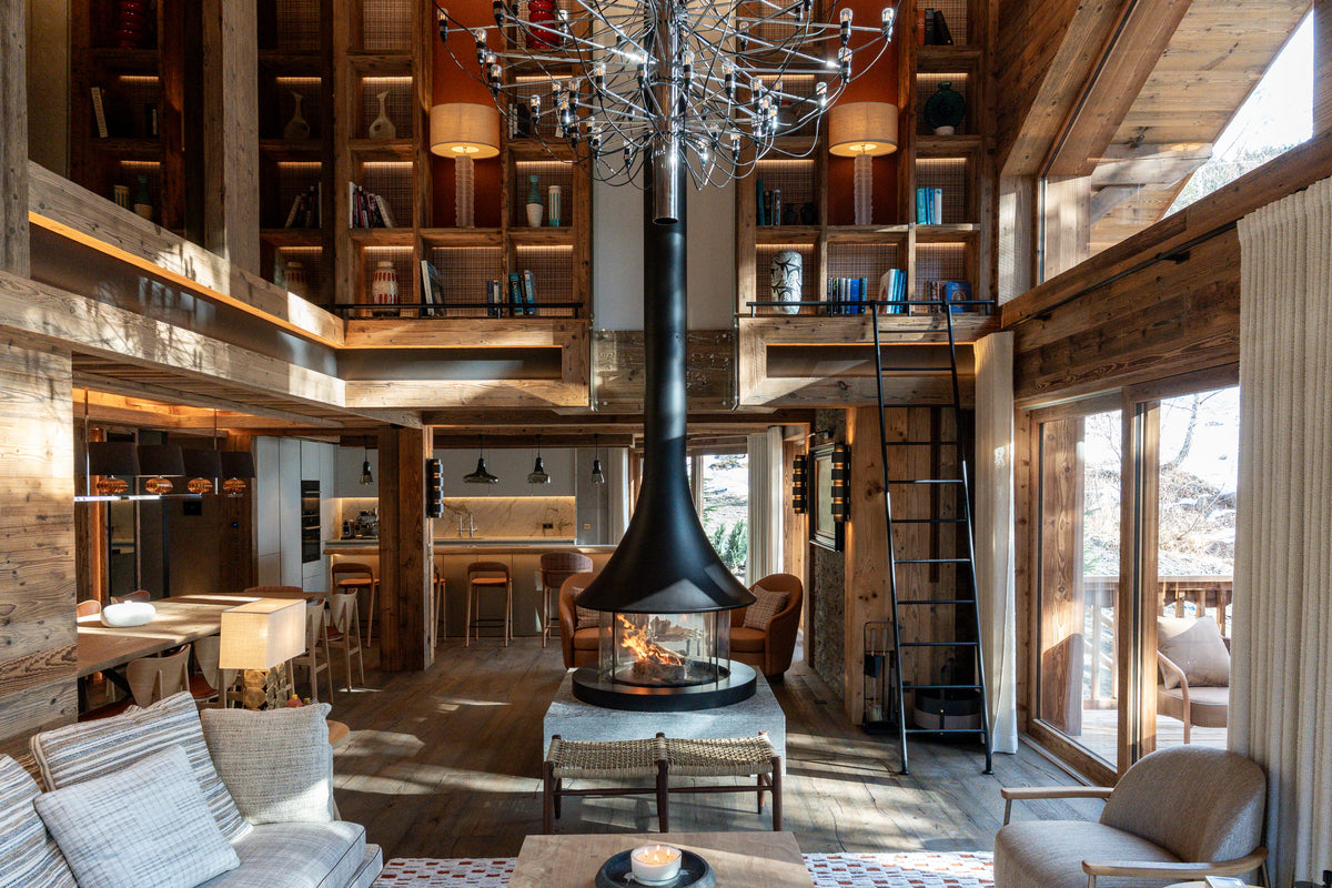 A high-ceilinged rustic lodge living room with a central black cone fireplace, tall wood bookshelves, large windows, and outdoor views.