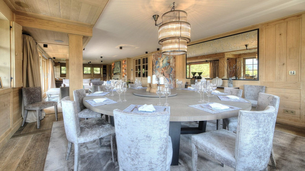 A large round dining table set with white tablecloths, wine glasses, and placemats, surrounded by gray upholstered chairs in a rustic wood-paneled dining room.