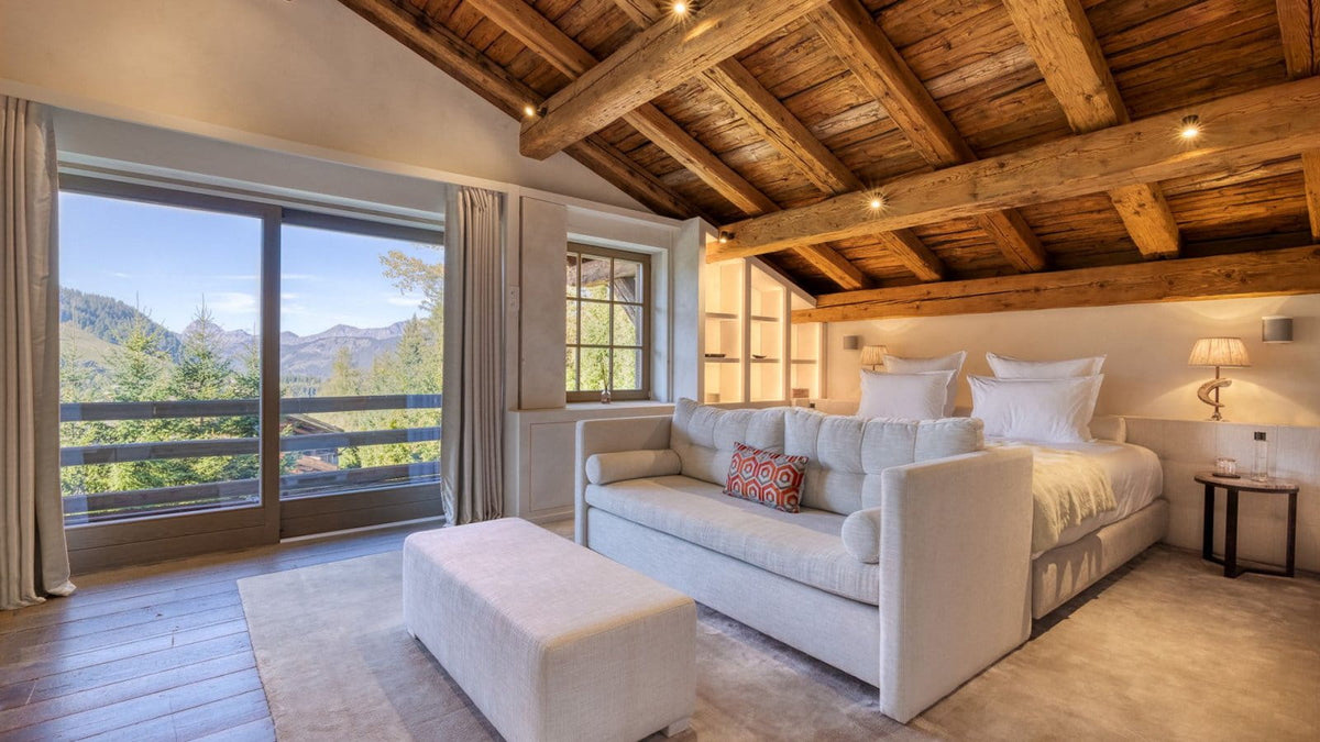 A luxury mountain chalet bedroom featuring a large off-white sofa, a matching ottoman, a wooden bed frame, and a rustic wood-beamed ceiling, with a large window showcasing a scenic forested mountain view.