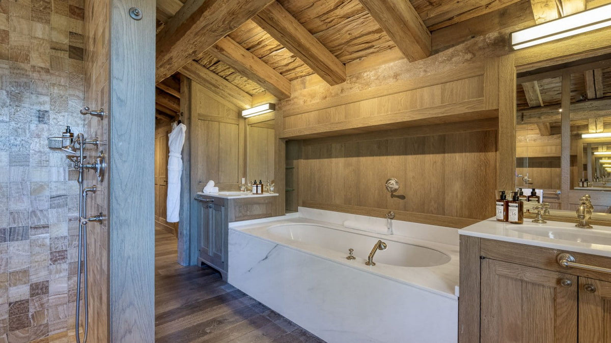 Luxury rustic bathroom featuring a large white freestanding bathtub, wooden vanity cabinets, a stone-tiled shower area, and a high wood-beamed ceiling.