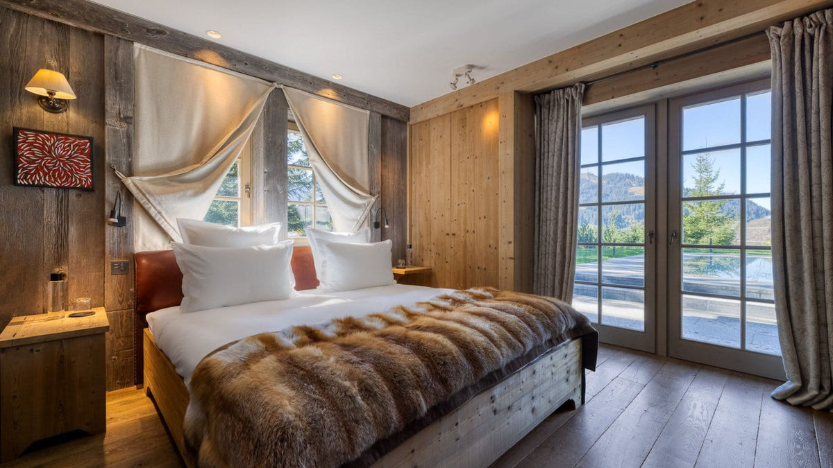 A rustic mountain chalet bedroom featuring a large bed with white linens and a brown fur throw, set against wood-paneled walls with draped curtains and a glass door overlooking a pool.