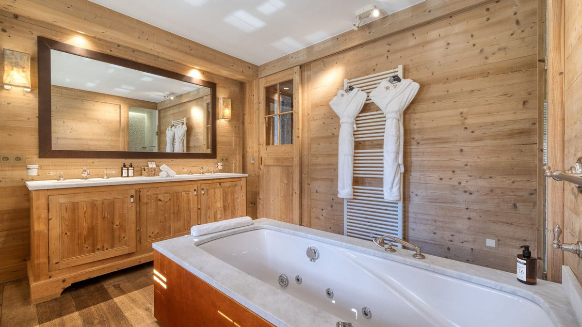 Luxury spa bathroom featuring a large white hot spring jacuzzi tub with a warm wood surround, set against rustic wood-paneled walls. A matching wooden double vanity with a large mirror and white marble countertop is on the left, and a white towel draped over a heated towel rail hangs on the right.