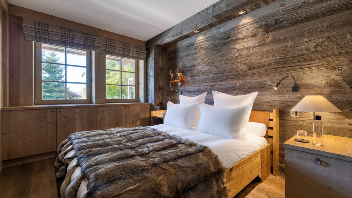 A rustic mountain-style bedroom featuring a wooden bed frame, white linens, a plush gray fur throw blanket, a matching wood headboard, and a large wood-paneled wall with built-in wall lamps.
