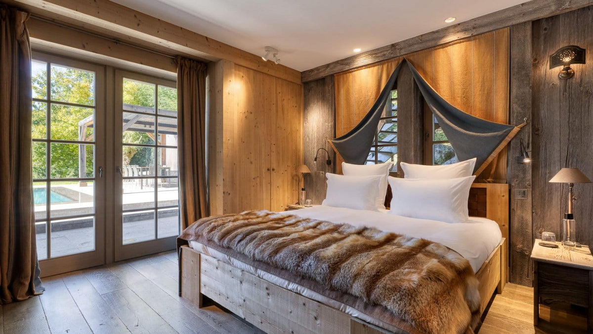 A rustic mountain-style bedroom with a large bed dressed in white linens and a brown fur throw, set against wood-paneled walls with a dark canopy. Large glass doors overlook a pool area, and bedside tables with lamps sit on both sides.