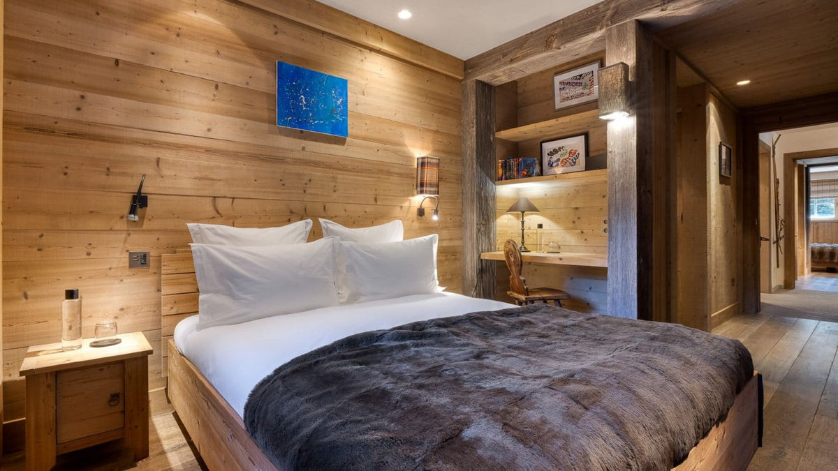 A cozy, rustic bedroom with a wooden bed dressed in white linens and a dark gray faux-fur throw, set against a wood-paneled wall with a small blue artwork and built-in shelving.