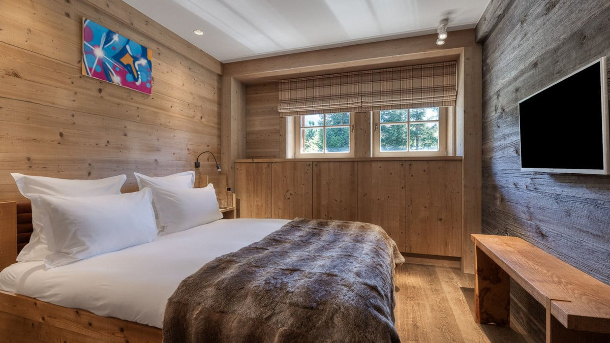 A cozy bedroom interior with a neatly made bed, white bedding, and a brown textured throw blanket in a rustic wood-paneled room. A wall-mounted TV, wooden bench, window with blinds, and colorful wall art are visible.