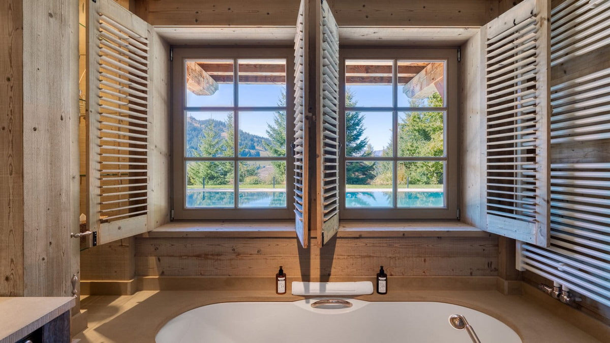 A rustic wooden bathroom interior with an oval white bathtub in the foreground, large shuttered windows, and a scenic mountain lake view outside.