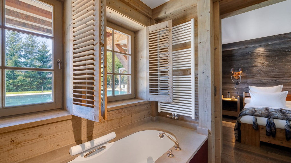 A luxury bathroom interior with a white freestanding bathtub, white shutters, and light wood walls, with an open doorway revealing a bedroom with a bed and dark wood headboard.