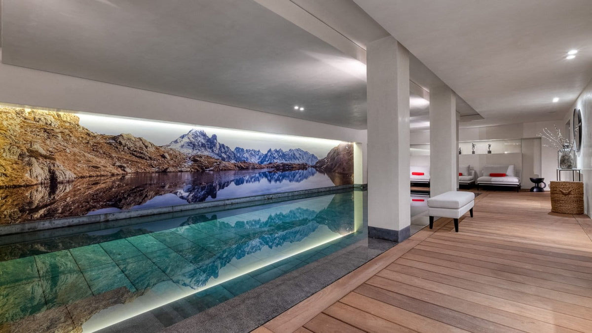 An indoor swimming pool with a large wall mural depicting a mountain lake scene, set in a modern room with wood flooring and white columns.