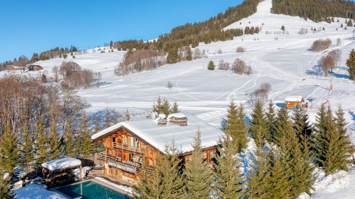 A snowy mountain landscape with a wooden chalet, evergreen trees, ski slopes, and a clear blue sky.