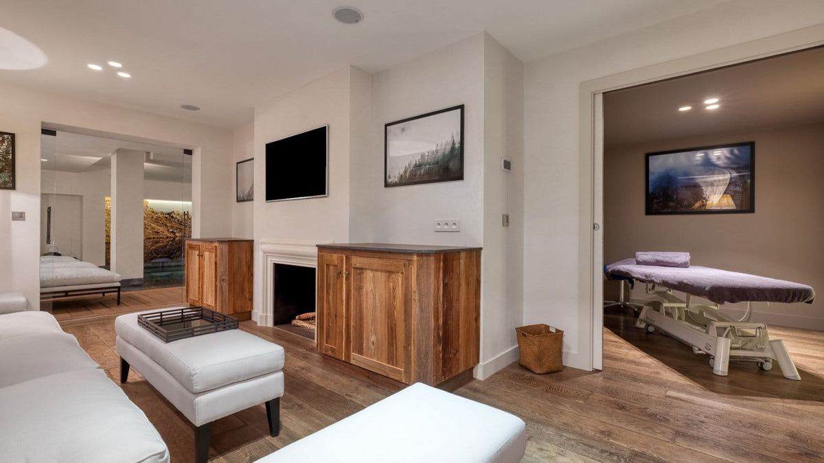 A modern interior room with hardwood floors, white upholstered seating, wall-mounted framed art, a fireplace with wood cabinets, and an open doorway revealing a treatment table in an adjacent room.