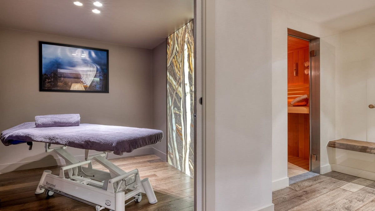 A spa treatment room featuring a white adjustable massage table with a purple blanket and pillow, a framed landscape print on the wall, a decorative glass partition, and an open doorway revealing a wooden sauna interior.