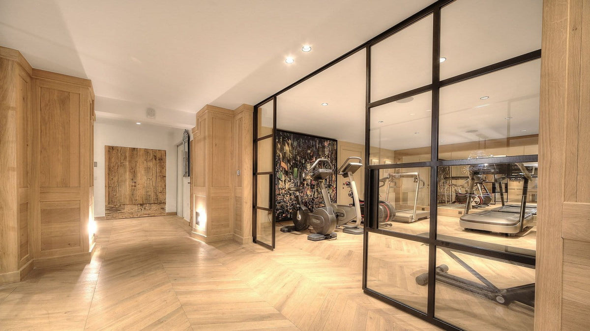 A modern home gym interior with light wood flooring, wooden cabinetry, and a glass-walled enclosure containing exercise equipment including a treadmill and stationary.