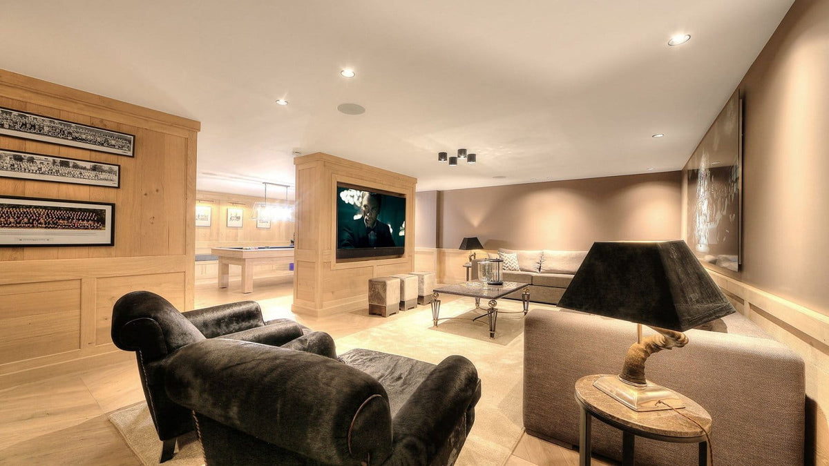 A modern indoor lounge or game room with dark velvet armchairs, a beige sofa, a side table with a black lamp, wood-paneled walls, framed artwork, and a television mounted on a wall.