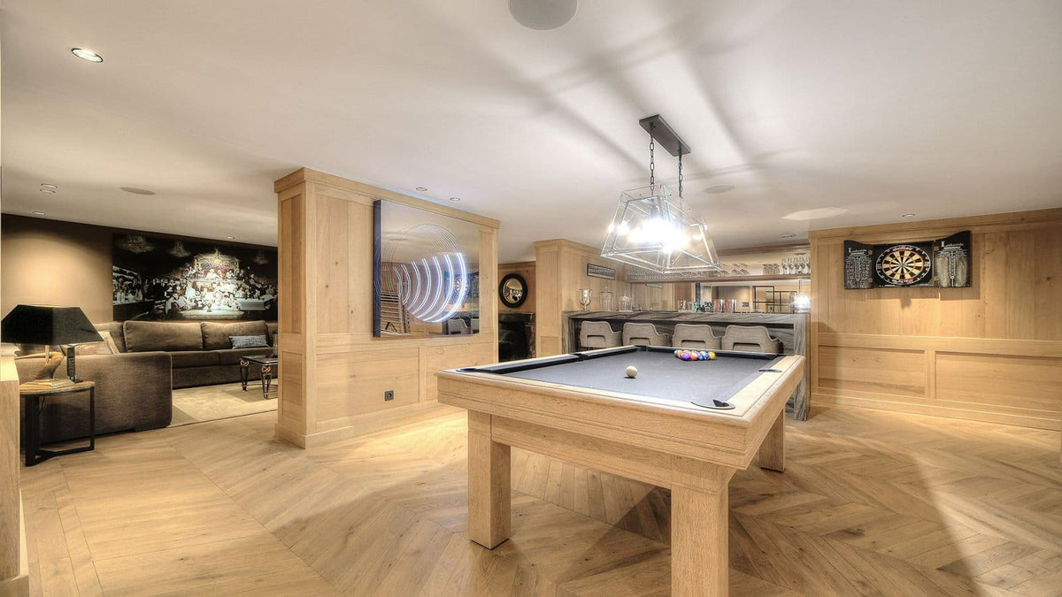 A large wooden game room with a pool table in the foreground, wood-paneled walls, a hanging pendant light, and a bar area in the background.