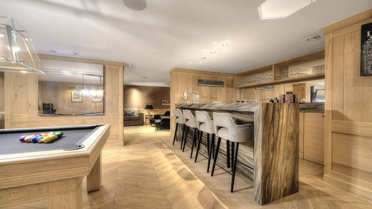 A modern indoor bar and pool area with light wood cabinetry, a wood-look bar counter, four gray upholstered bar stools, a pool table in the foreground, and a bright open ceiling with recessed lighting.