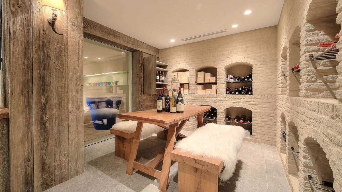 A rustic indoor dining or wine cellar nook with a wooden table and two white faux-fur benches, set against a light stone brick wall with built-in wine shelving and bottles.