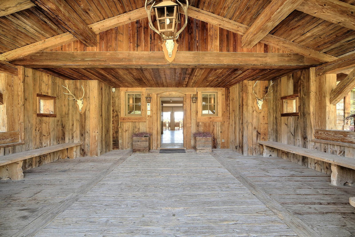A rustic wooden lodge interior with a high vaulted ceiling, exposed beams, long wooden benches on both sides, a central doorway, mounted deer heads on the walls, and a hanging lantern light fixture.